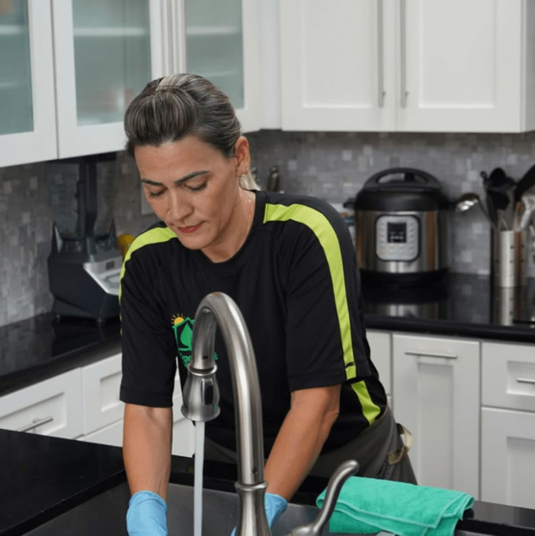 A professional cleaner wearing a black uniform with green stripes and blue gloves is washing a cloth under running water in a modern kitchen. The countertops are clean and organized, reflecting the consistency and care of a recurring cleaning service. 